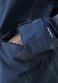 Tmavě modrá bunda se zapínací kapsou na zip a suchým zipem na manžetě, s texturovaným vzorem a nápisem "DRYVENT" na rukávu.