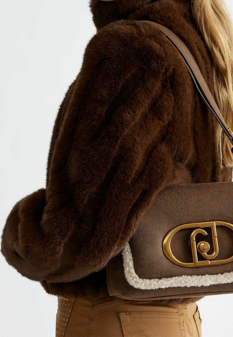 Femme portant une veste en fourrure marron foncé et un sac à main en cuir marron avec une fermeture dorée et une bordure en shearling blanc sur l'épaule.