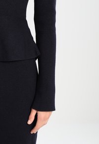 Robe bleu marine foncé ajustée à manches longues avec un détail péplum à la taille. Texture côtelée sur les manches et le péplum, lisse sur le corps.