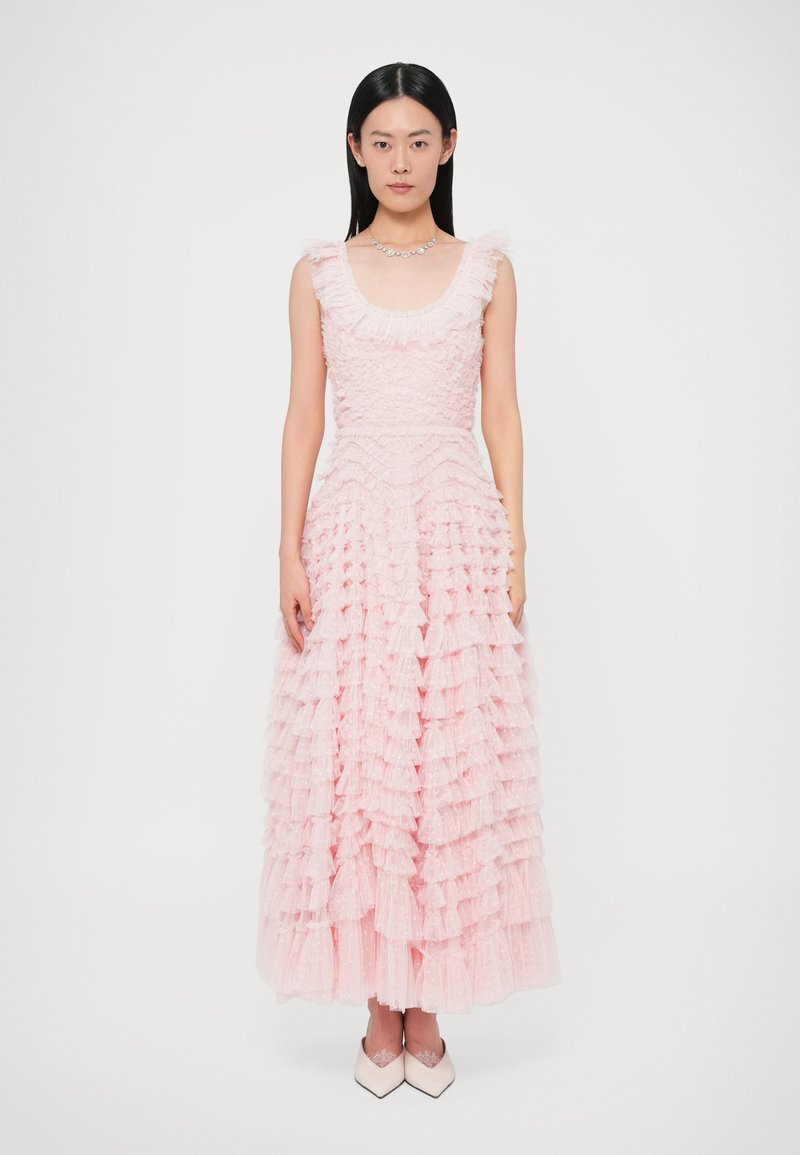 Ráncolt rózsaszín ruha alakra szabott felsőrésszel, mélyen bevágott nyakkivágással és padlószőnyegig érő, réteges fodrokkal díszített szoknyával. Fehér, hegyes orrú cipővel párosítva.