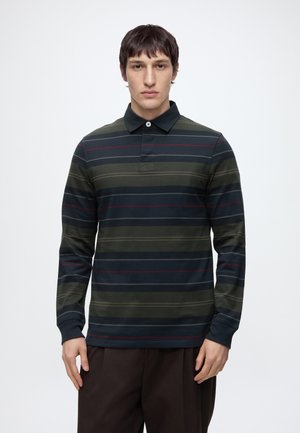 Jeune homme portant un polo sombre à manches longues avec des rayures horizontales vertes, bleu marine et rouges, debout devant un fond blanc uni.