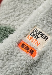 Fleece-stof i lysegrå med en blød, tekstureret overflade. Har et vævet mærke med "SUPER DRY MNTN" i sort og orange accenter.