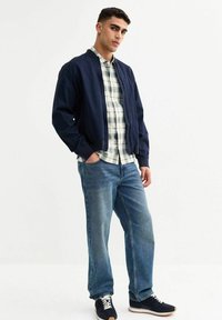 Giacca bombardiere navy su una camicia a quadri chiari, abbinata a jeans blu e sneakers scure. Caratterizzata da una vestibilità rilassata e una superficie morbida.