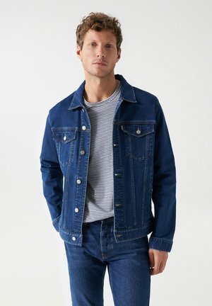 Chaqueta de denim azul oscuro con un cuello clásico, bolsillos frontales y botones plateados, llevada sobre una camiseta de rayas y jeans a juego.