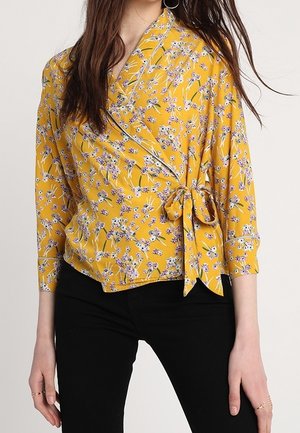 Woman wearing a yellow floral wrap blouse with 3/4 sleeves tied at the side, paired with black pants against a neutral background.