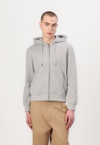 Felpa grigia con zip, tasca frontale, cappuccio con lacci e un piccolo logo sul lato sinistro del petto. Abbinata a pantaloni khaki.