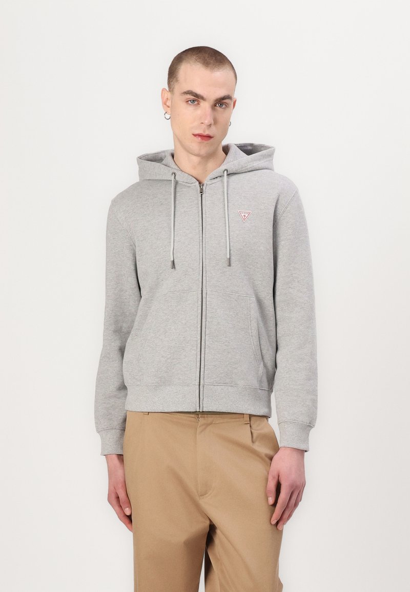 Felpa grigia con zip, tasca frontale, cappuccio con lacci e un piccolo logo sul lato sinistro del petto. Abbinata a pantaloni khaki.