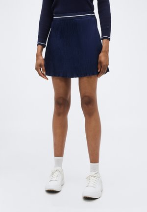 Osoba ve tmavě modré plisované sukni, tmavě modrém dlouhém tričku s bílým lemováním, bílých ponožkách a bílých teniskách stojí před jednobarevným pozadím.