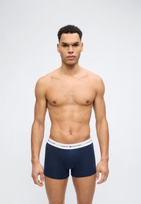 Μυώδης άνδρας με κοντά, σγουρά μαλλιά στέκεται με το πρόσωπο προς τα εμπρός, φορώντας μπλε σκούρα μποξεράκια Tommy Hilfiger με λευκή μέση.