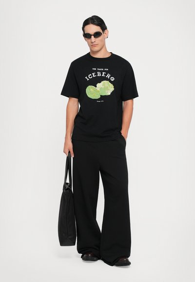 Black cotton t-shirt featuring a green graphic of lettuce, paired with loose black pants and a black tote bag. The outfit has a casual design.