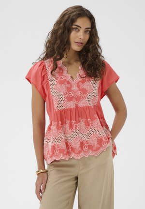Kobieta w koralowej bluzce z koronkowym kwiatowym haftem i beżowych spodniach, stoi z jedną ręką w kieszeni na białym tle.
