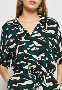 La chemise présente un motif abstrait de vagues en noir, blanc et vert, avec un col, des manches courtes et un détail de taille nouée.
