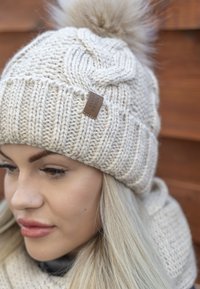 Világos bézs kötött sapka műszőrme pom-pom-mal és bőr címkével. Texturált mintákkal és bordázott kialakítással rendelkezik a kényelem érdekében.