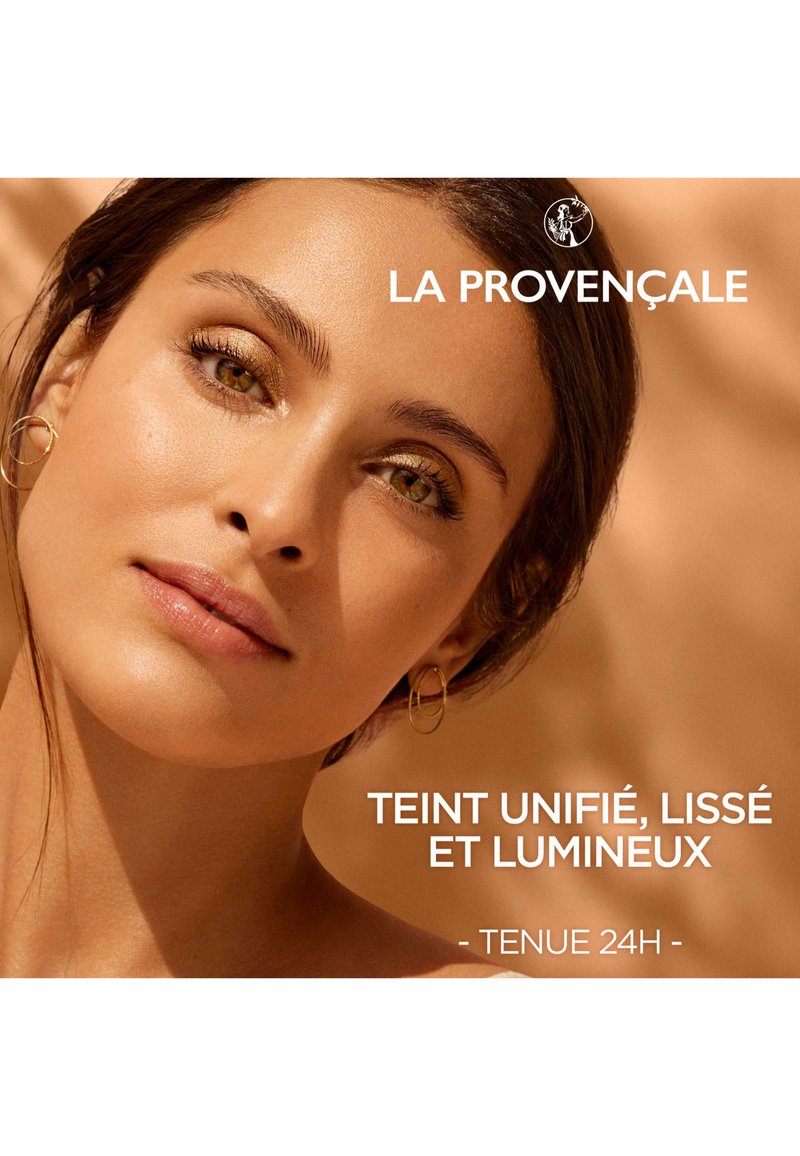 Gros plan sur un visage avec un teint chaleureux, une texture lisse et des traits bien définis. Du fard à paupières doré accentue les yeux verts. De petites boucles d'oreilles en forme de cercle sont visibles.