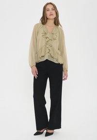 Blusa beige a maniche lunghe con rouches lungo il davanti, abbinata a pantaloni neri a gamba larga a vita alta e scarpe con tacco a punta nere.