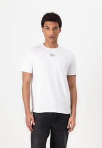 Hvid bomulds T-shirt med korte ærmer, rund hals og et lille gråt logo på brystet. Sammen med mørke denim jeans.