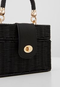 Sac à main tissé noir avec une sangle texturée, une fermeture à fermoir tournant doré et des attaches de poignées en corde noire torsadée sur un fond clair.