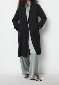 Černý dvouřadý trenčkot s dlouhými rukávy, postranními kapsami a volným střihem, nošený přes světle zelenou košili na knoflíky a široké kalhoty.