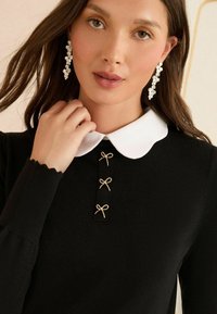 Melnā džemperis ar baltu apkakli, kas rotāta ar trim zelta loku akcentiem apakšā. Garas piedurknes ar viļņotām maliņām.