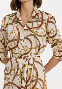 Blusa stampata in tessuto color crema chiaro con motivi di catene e cinture dorate. Maniche lunghe, chiusura con bottoni frontale e dettaglio vita annodato.