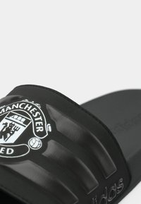 Melnas slaidu sandales ar teksturētu siksnu, uz kuras redzams balts "Manchester United" logo. Pēdas daļa ir gluda ar reljefā iespiestu zīmola nosaukumu.