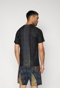 Μαύρο αθλητικό μπλουζάκι με υφή υφάσματος, με χρυσές λεπτομέρειες και κάθετη ρίγα. Συνδυάζεται με σορτς με σχέδιο σε ναυτικό μπλε και χρυσό.