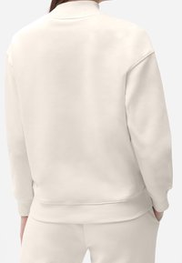 Sudadera de color beige claro con cuello alto, puños y dobladillo de canalé. Fabricada con una tela suave, presenta un diseño sencillo y un ajuste relajado.