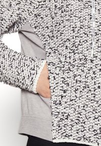 Huvtröja med ett texturerat, vitt och svart mönstrat yttertyg och ett slätt grått innerfoder. Har en framficka och dragsko.