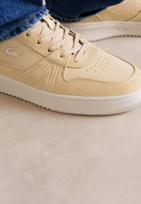 Bežové tenisky s textúrovanou kožou, okrúhlou špičkou, plochou bielou podrážkou a klasickými šnúrkami. S logom na boku a perforovanými detailmi.