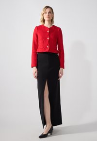 Veste courte rouge en tissu texturé, avec des boutons dorés et deux poches avant ; associée à une jupe maxi noire à fente haute et des talons noirs.