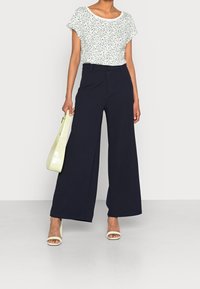 Vrouw in een witte bloemenprint met korte mouwen en hoge zwarte wijde broek, met een lichtgroene handtas in de hand, staand op een witte vloer.