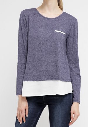Vrouw draagt een paarse top met lange mouwen, een witte gelaagde zoom en donkerblauwe jeans, staand tegen een effen achtergrond.