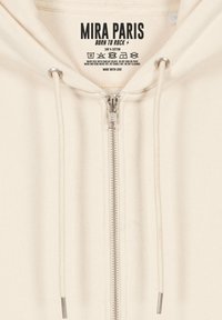 Felpa in cotone color crema con zip frontale, cordini e un'etichetta con la scritta "MIRA PARIS, BORN TO ROCK." Presenta hardware argentato.