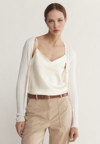 Vit ribbad cardigan över en krämfärgad satin-camisole, matchad med beiga byxor och ett brunt bälte. Släta texturer och subtila färger.