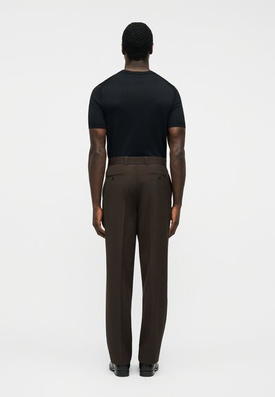 Άνδρας που φοράει μαύρο κοντομάνικο πουκάμισο και σκούρο καφέ ραμμένα παντελόνια, στέκεται με την πλάτη προς τα εμπρός σε απλό φωτεινό φόντο.