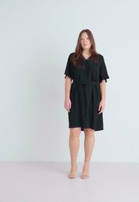 Robe noire à manches courtes avec décolleté en V, détail de ceinture à nouer et coupe décontractée. Tissu lisse avec un ourlet au genou. Assortie à des sandales transparentes.
