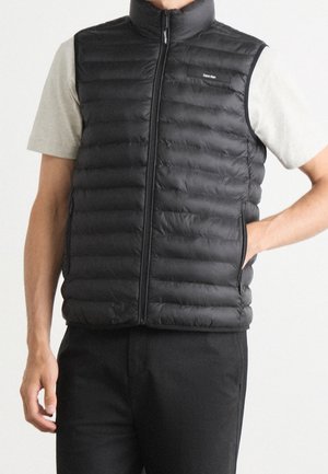 Mand iført en sort quiltet ærmeløs vest over en lys kortærmet skjorte, med den ene hånd i lommen og sorte bukser.