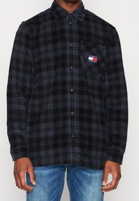 Uomo che indossa una camicia a quadri blu scuro e nera con il logo Tommy Jeans sulla tasca del petto e jeans azzurri chiari.