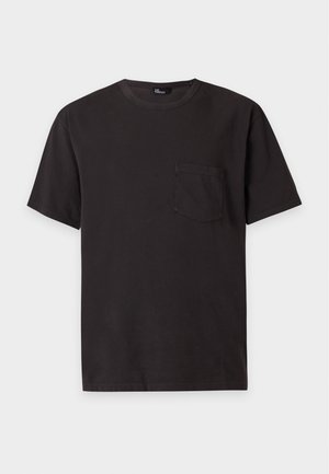 Jeune homme debout portant un t-shirt noir avec poche, un jean bleu clair et des chaussures en cuir noires sur un fond blanc uni.