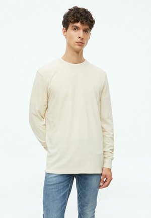 Jonge man met bruin krullend haar, gekleed in een beige overhemd met lange mouwen en blauwe spijkerbroek, staande tegen een effen witte achtergrond.