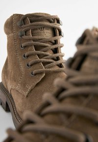 Bottes chevilles en daim marron avec œillets en métal, lacets ronds et semelle en caoutchouc robuste. Présente des coutures visibles et une étiquette de marque sur le côté.