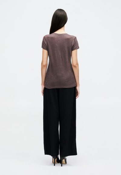 Femme aux longs cheveux bruns portant une chemise marron moulante à manches courtes, un pantalon large noir et des talons hauts noirs, debout dos à la caméra.