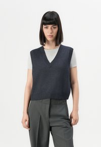 Chaleco de punto azul marino con escote en V, con ribete en el dobladillo, usado sobre una camiseta de color gris claro, combinado con pantalones gris de cintura alta.