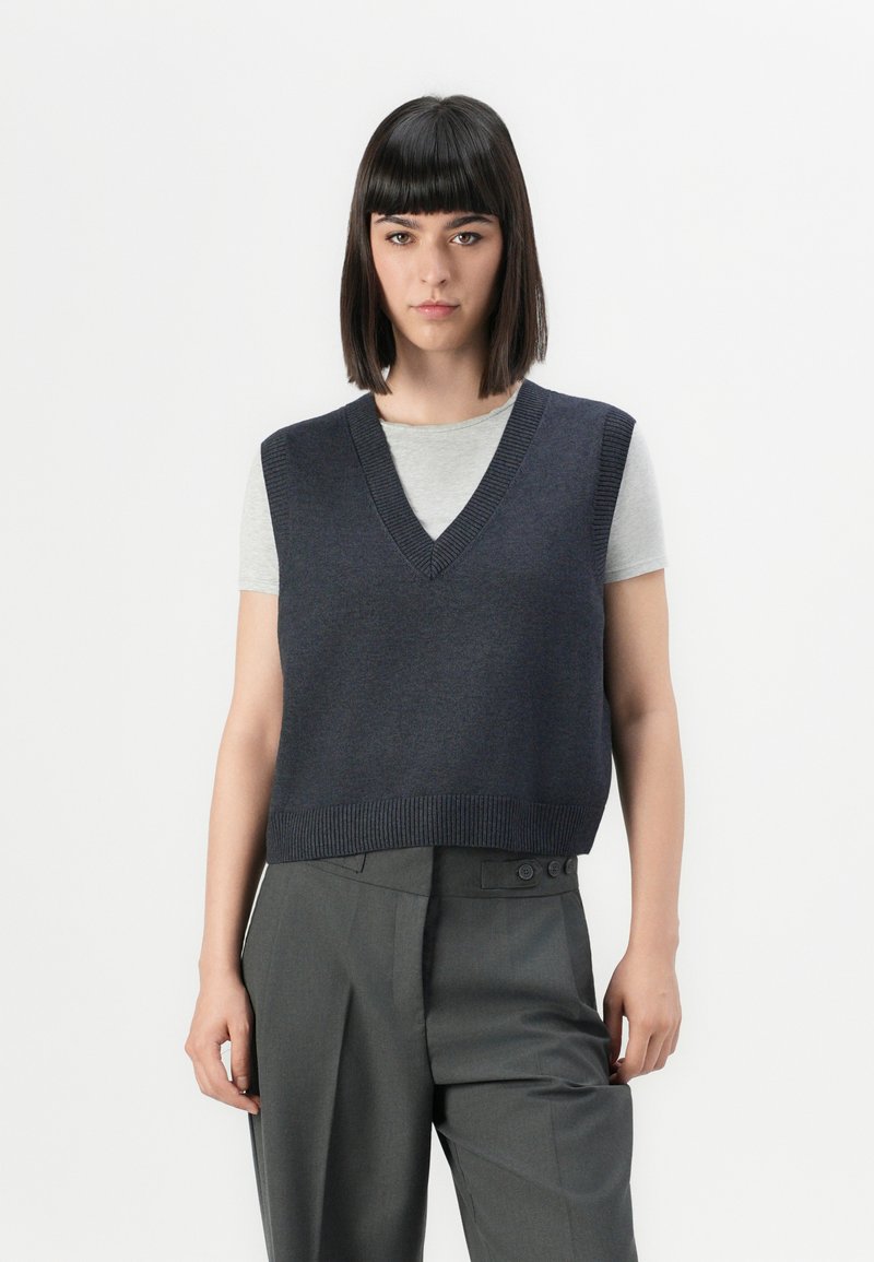 Chaleco de punto azul marino con escote en V, con ribete en el dobladillo, usado sobre una camiseta de color gris claro, combinado con pantalones gris de cintura alta.