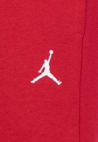 Sarkanais kokvilnas audums ar balto izsistā Jumpman logotipu kreisajā pusē. Audumam ir gluda tekstūra, un dizains ir minimālistisks.