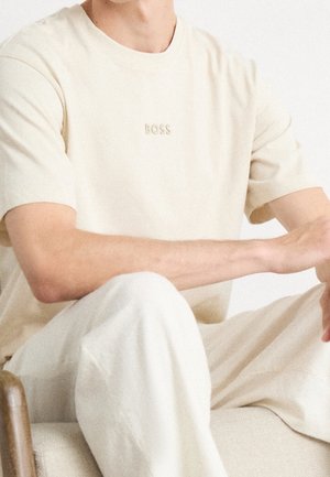 Beżowa bawełniana koszulka z krótkim rękawem oraz okrągłym dekoltem. Z przodu znajduje się haftowane logo "BOSS" na piersi, zestawiona z jasnymi spodniami.