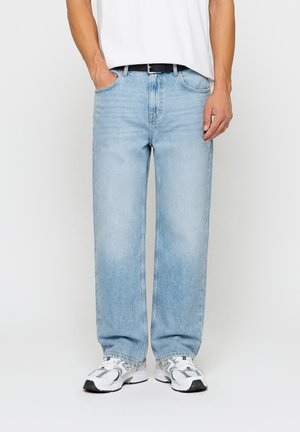Person iført lyseblå straight-leg jeans, hvide sneakers, en hvid skjorte stukket ind og et sort bælte, stående foran en hvid baggrund.