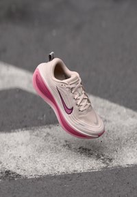Nike futócipő, világos bézs hálós felsőrész, rózsaszín középtalp, lila swoosh logó, lekerekített orr, struktúrált dizájn, fehér jelölés felett lebeg egy sötét felületen.