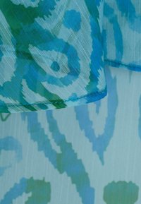 Gros plan sur un tissu translucide bleu et vert à motifs, avec des designs abstraits circulaires et ondulés, montrant des coutures le long de l'ourlet.