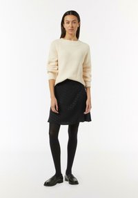 Cremefarbener Strickpullover mit langen Ärmeln, kombiniert mit einem schwarzen, strukturierten Rock. Schwarze Strumpfhosen und Loafers runden das Outfit ab.